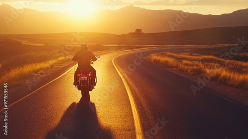 A motorcyclist rides down a winding road at sunset, casting a long shadow on the asphalt. The sun is setting behind the rider and creating a warm glow over the landscape.