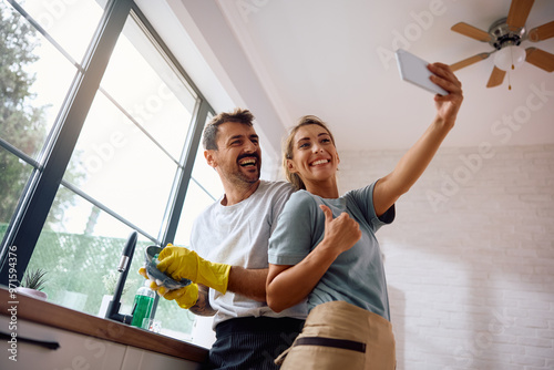 Happy woman taking selfie with her boyfriend who is doing the dishes in kitchen.