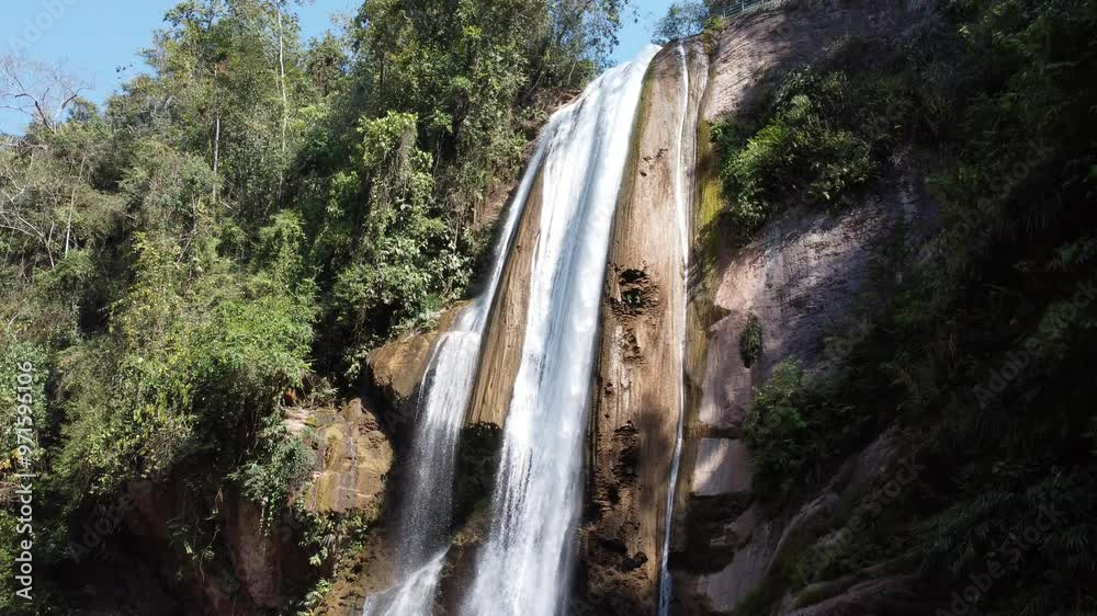 Camera pans to show a waterfall cascading down a rocky cliffside surrounded by lush greenery Camera pans to show a waterfall cascading down a rocky cliffside surrounded by lush greenery