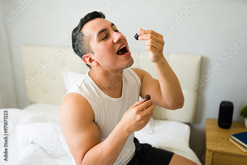 Foto Hispanic man in his bedroom, taking his daily cbd oil dose with a dropper, promo