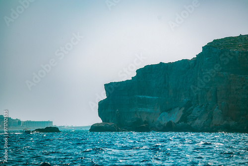 Rocky cliff overlooking the ocean on a misty day, with distant buildings and a boat visible near the horizon
