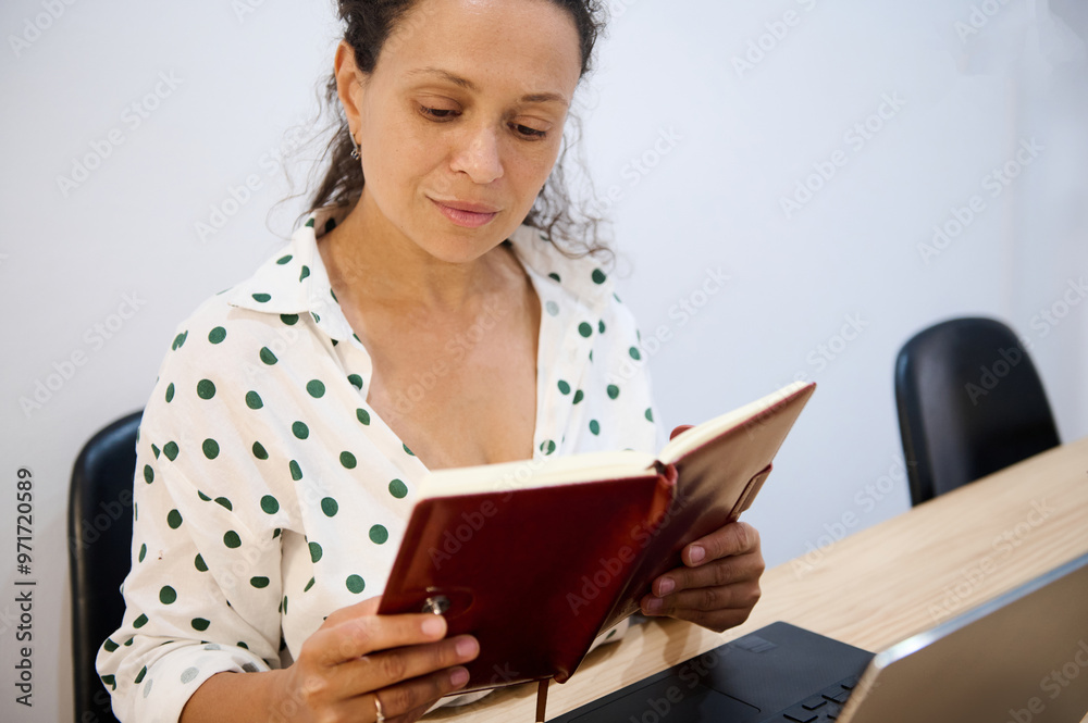 Woman in polka dot blouse reading a book while working at desk with a laptop in a quiet office setting Woman in polka dot blouse reading a book while working at desk with a laptop in a quiet office setting