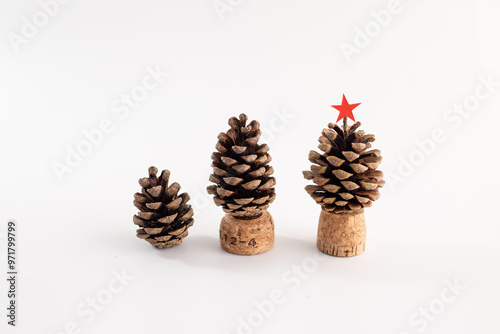 Three pine cones on white surface, mounted on cork bases, resembling tiny trees. Largest cone topped with red star, showcasing festive DIY craft with natural materials, perfect for holiday decor