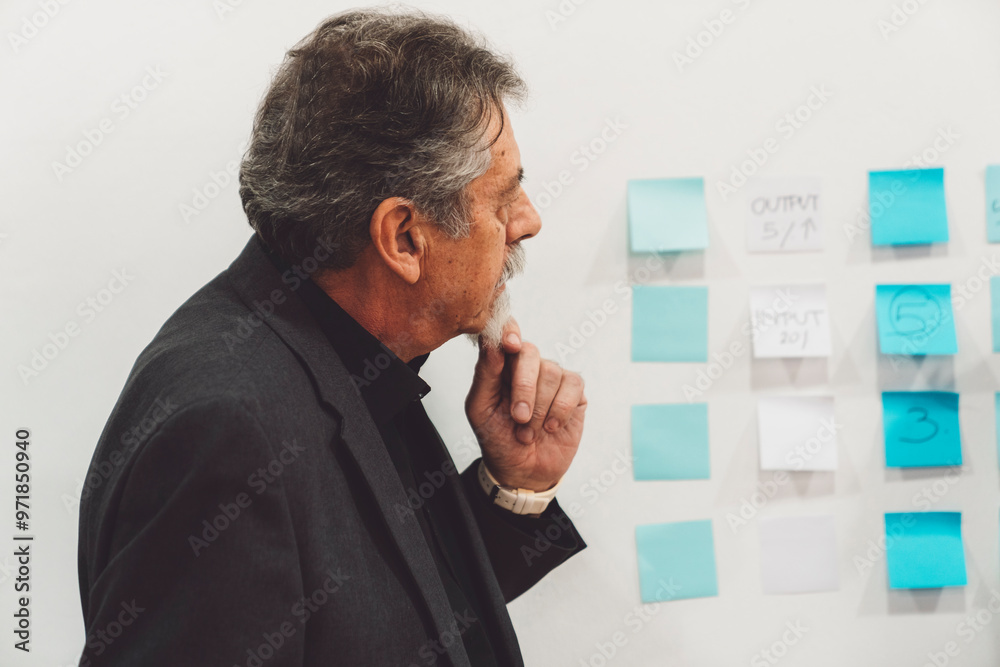 Senior man holding for his chin, thinking, looking at sticky notes on the wall Senior man holding for his chin, thinking, looking at sticky notes on the wall