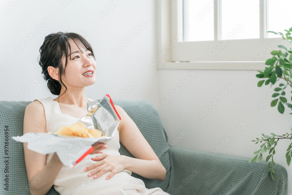 自宅のソファでポテトチップスを食べる若いアジア人女性（暴飲暴食・間食・高カロリー）
