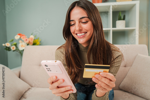 Fotografie Young caucasian woman using a smart phone to make an online purchase with a credit card sitting at home sofa
