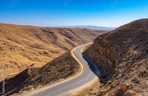 A long, winding road cutting through a desert landscape, with rocky hills and a clear blue sky stretching out into the horizon