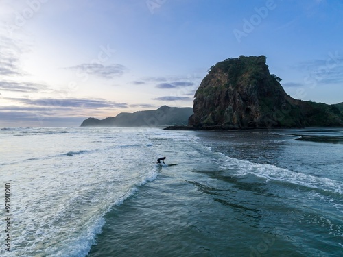 Surfer exiting the water on the beach at sunset. Lion Rock is in the background, Piha, Auckland, New Zealand. photo