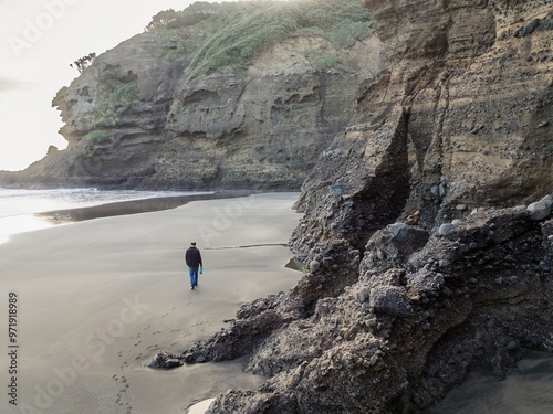 Man walking along a sandy shoreline next to cliffs in Piha, Auckland, New Zealand.