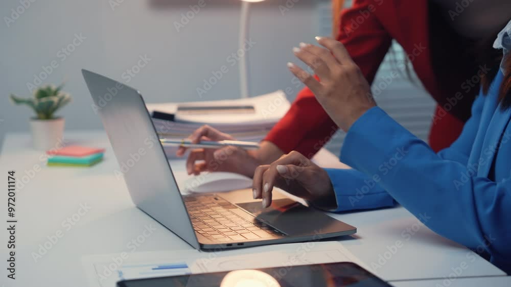 Two businesswomen in suits collaborate at a desk, analyzing data on a laptop and digital tablet in a corporate office setting, focusing on their project and meeting deadlines