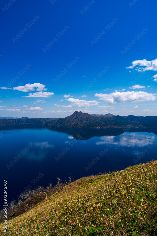 雲の浮かぶ青空を反射する山間の湖。日本の北海道の摩周湖は美しい自然の景色が観光お客に人気の名所。雄大で迫力のあるカルデラ湖の風景が魅力。 雲の浮かぶ青空を反射する山間の湖。日本の北海道の摩周湖は美しい自然の景色が観光お客に人気の名所。雄大で迫力のあるカルデラ湖の風景が魅力。