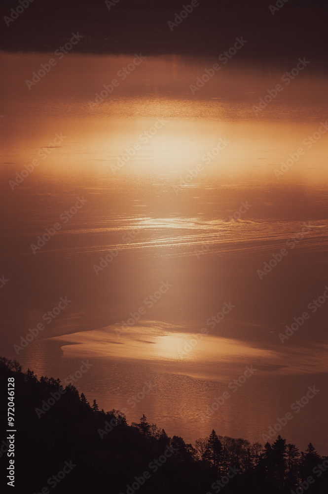 Obraz premium 広大な湖面に太陽の光が反射し、水面全体が黄金色に輝いている。遠方にはシルエットになった山々が連なり、雄大な自然の風景が広がる。金色の光が湖を照らし、神秘的な雰囲気を醸し出す。