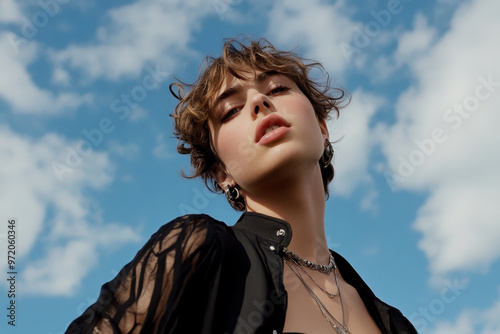 A confident individual with short, curly hair poses against bright blue sky, showcasing stylish outfit and accessories. image captures sense of modern gender fluidity and androgyny, reflecting contemp photo