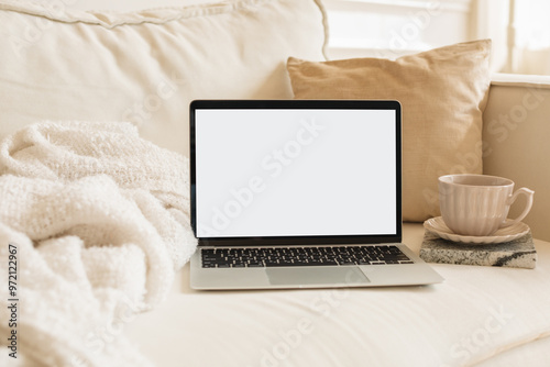A laptop with a white screen mock-up and a beautiful coffee cup on a cozy light sofa, surrounded by pillows and a warm blanket in a modern living room.