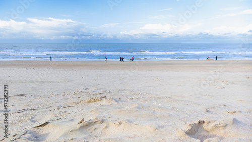 A serene coastal scene showing a wide sandy beach during low tide with a few people strolling under a cloudy sky in autumn.