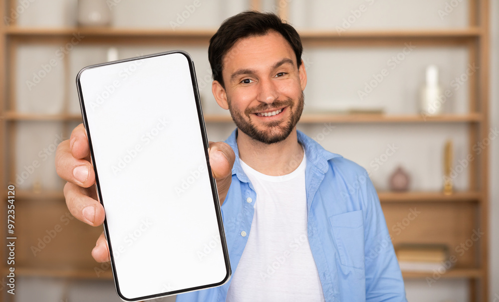 A man stands in a bright, inviting room, smiling as he holds out a blank smartphone. The background features shelf decor, creating a warm atmosphere A man stands in a bright, inviting room, smiling as he holds out a blank smartphone. The background features shelf decor, creating a warm atmosphere