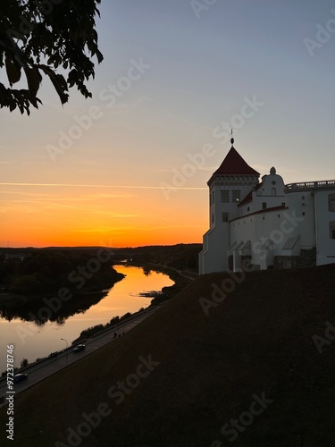 view of white medieval castle with red roofs on tower near river on cliff in summer hot sunset