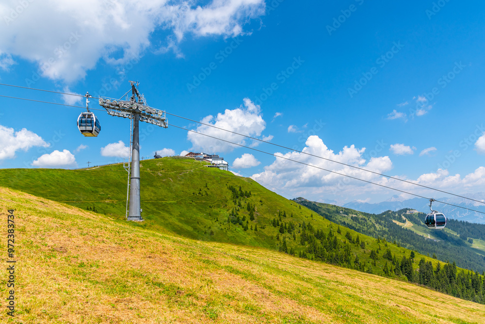 Experience the stunning views from the Schmittenhohe cableway in the Austrian Alps, gliding over lush green hills on a clear day filled with fluffy clouds.