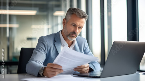 Photo of a middle-aged businessman sitting at his desk in an office, looking over papers with a thoughtful expression while using a laptop computer. 