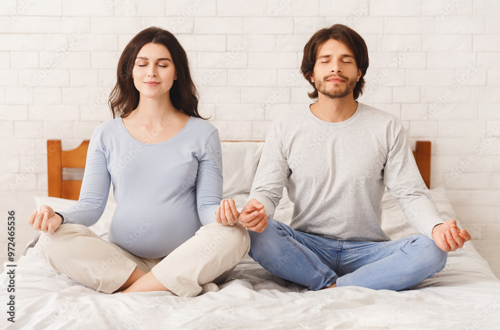 Prenatal yoga. Pregnant woman and her husband doing breathing exercises, sitting in lotus position and meditating together on bed at home, free space Prenatal yoga. Pregnant woman and her husband doing breathing exercises, sitting in lotus position and meditating together on bed at home, free space