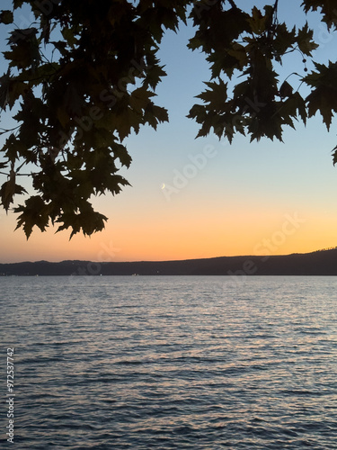 A crescent moon rises above Bracciano Lake at dusk, framed by tree leaves, in Trevignano Romano, Italy, with calm waters and a peaceful sky.