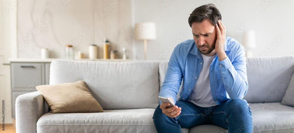 A man appears anxious on a couch in a cozy living room, holding his smartphone and grimacing. He seems overwhelmed by whatever he is reading or viewing on the screen. A man appears anxious on a couch in a cozy living room, holding his smartphone and grimacing. He seems overwhelmed by whatever he is reading or viewing on the screen.