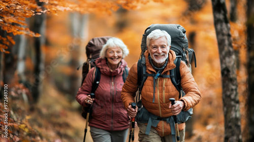 An energetic older couple strolls through a forest filled with vibrant autumn leaves, smiling as they wear hiking gear and carry walking sticks.