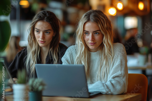 Two young women engaged in focused work on laptops while seated in a modern café, capturing a moment of collaboration and creativity in a bustling environment