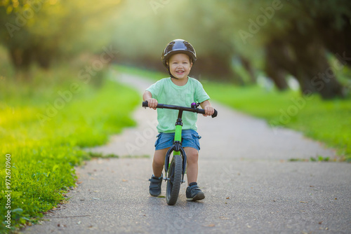 Happy smiling little boy with helmet running and riding on first bike without pedals on sidewalk at city park in warm summer day. Cute 2 years old toddler. Front view. Learning to keep balance. photo