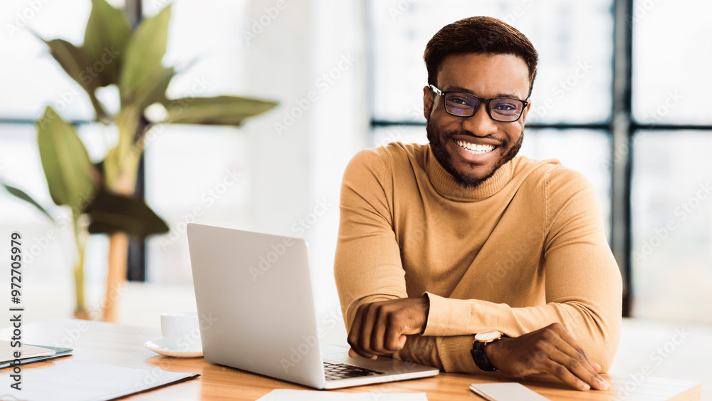 Success At Work. Smiling african guy sitting at desk, looking at camera, happy young professional. Panorama