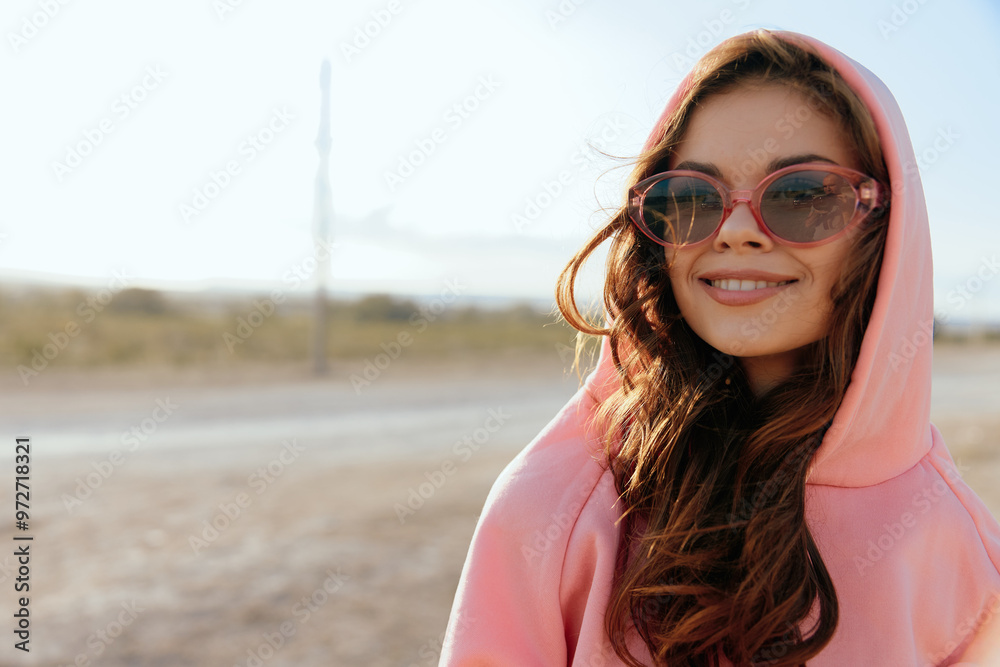 Woman in pink hoodie and sunglasses standing on dirt road with road in background on sunny day Woman in pink hoodie and sunglasses standing on dirt road with road in background on sunny day