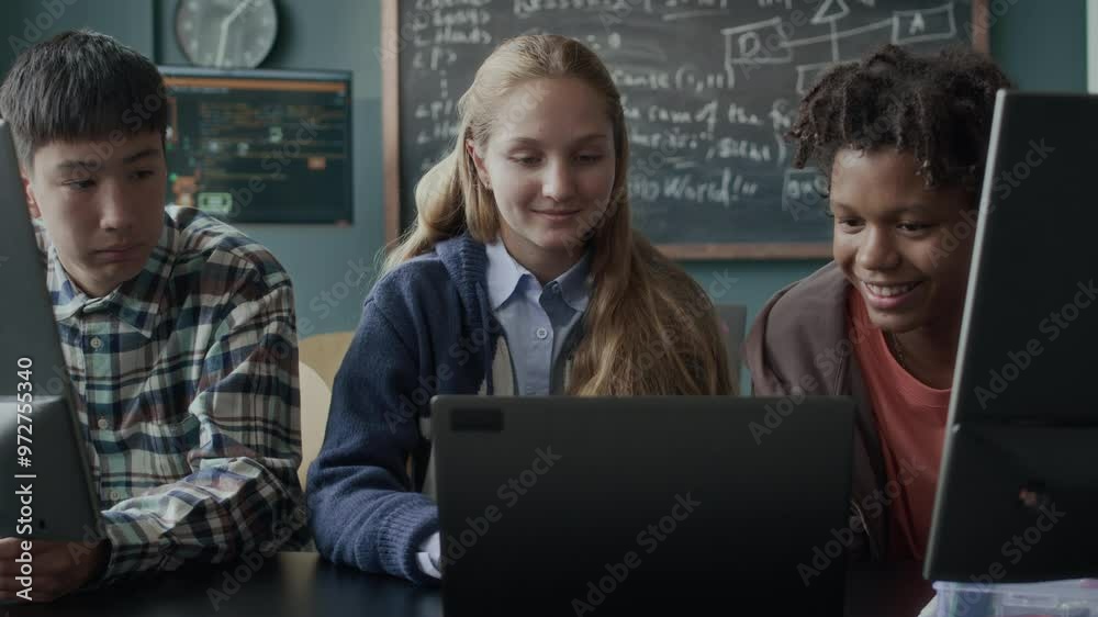 Medium shot of three multiethnic gen z school kids chatting while sitting in front of their computers at desk in modern programming class Medium shot of three multiethnic gen z school kids chatting while sitting in front of their computers at desk in modern programming class