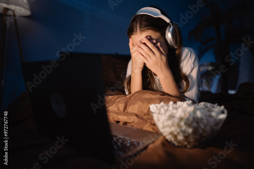 Obraz na plátne Teen girl eating popcorn and watching horror movie on laptop covering face with
