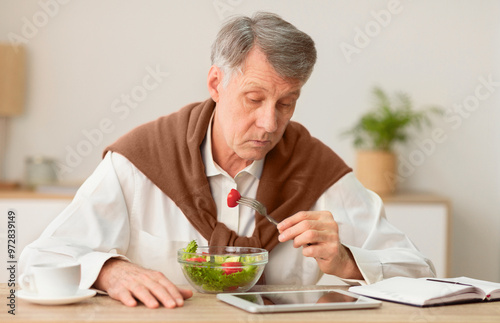 Senior Gentleman Eating Vegetable Salad Reading News On Tablet Computer Sitting At Workplace Indoor. Selective Focus