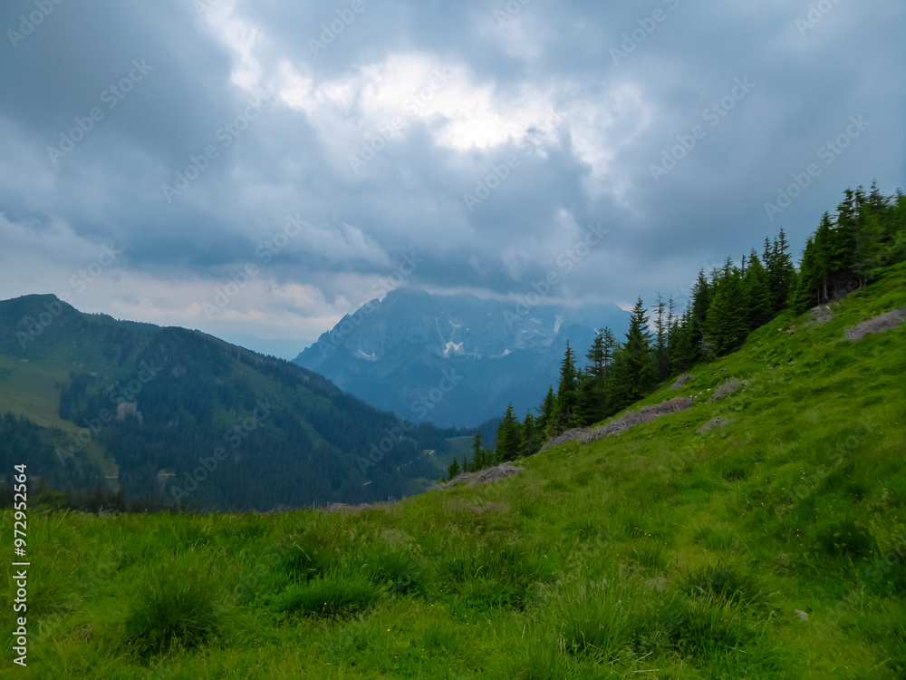 Obraz premium Hiking trail along alpine meadow with panoramic view of cloud covered mountain peaks in Eisenerz Alps, Styria, Austria. Tranquil atmosphere in Austrian Alps in spring. Outdoor trip on overcast day