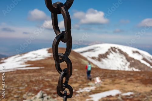 Close-up of a rusty chain hanging against snow covered alpine meadows and rolling hills to snow-capped mountain peak Lenzmoarkogel, Gleinalpe, Lavantal Alps, Styria, Austria. Woman hiking photo