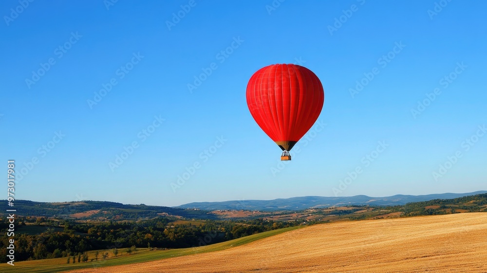 Fototapeta premium A bright red hot air balloon floating in a clear blue sky, casting a shadow on the landscape below.