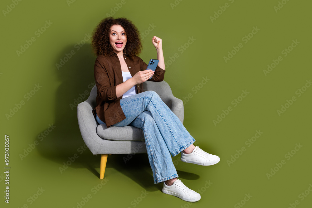 Full body portrait of nice young girl sit chair smart phone raise fist isolated on khaki color background Full body portrait of nice young girl sit chair smart phone raise fist isolated on khaki color background