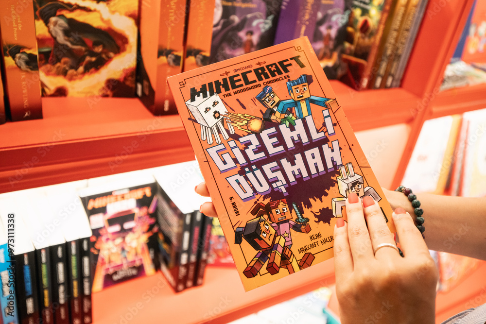 Fototapeta premium 09 July 2024, Istanbul, Turkey: A woman in a store, examining the cover of a Minecraft-themed novel, surrounded by merchandise