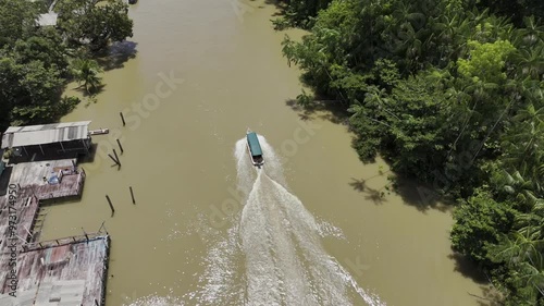 Drone pans up and follows green boat as it leaves forested river canal heading toward larger river photo