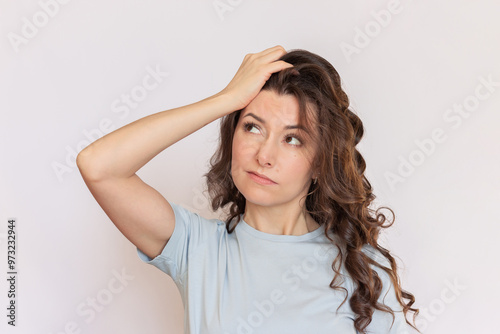 Young beautiful brunette woman with a curly hair and wearing a blue t-shirt looks thoughtfully to the side, puzzled by something, holding her hand on her head