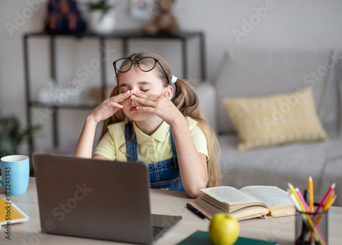 Tired schoolgirl rubbing her dry irritated eyes while studying online on laptop at home, copy space. Teen female student exhausted from looking at screen, doing assignments or communicating on web photo