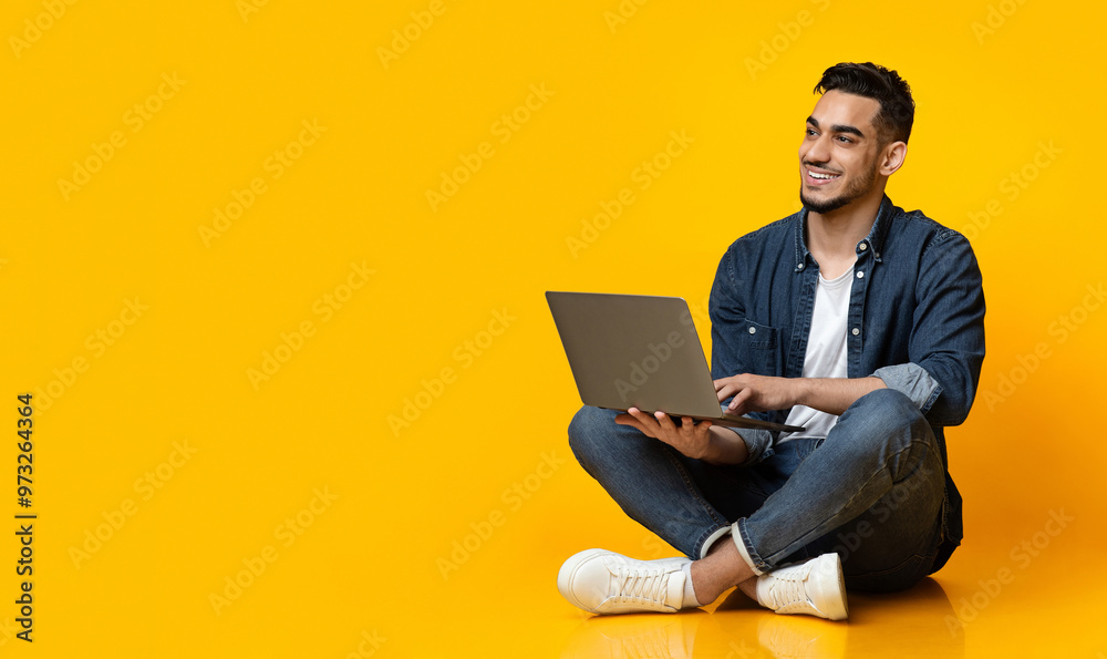 Happy arab young man sitting on floor, using new modern laptop, typing on keyboard and smiling over yellow studio background, looking at copy space for text or advertisement, panorama Happy arab young man sitting on floor, using new modern laptop, typing on keyboard and smiling over yellow studio background, looking at copy space for text or advertisement, panorama