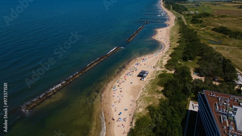 Drone captures Darłowo beach, golden sands, calm waters, sunbathers, nearby hotels, and a forest on the dunes.
