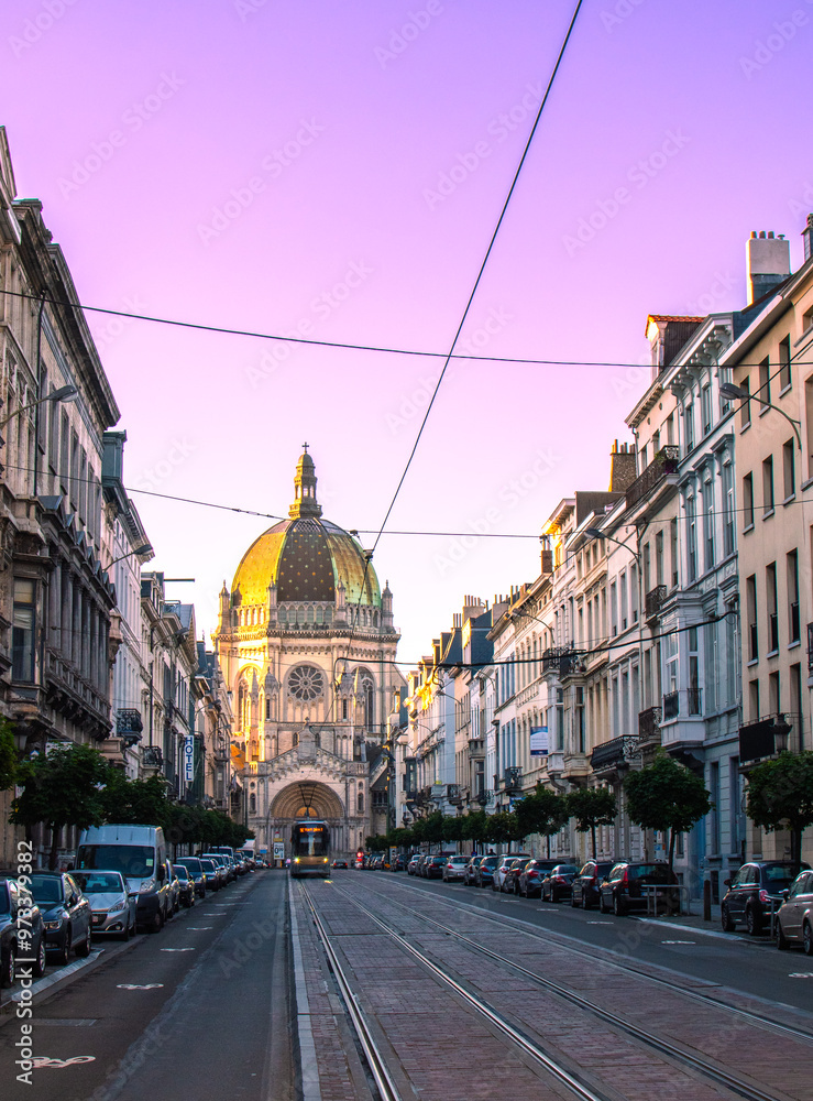 Naklejka premium A city street at sunset with buildings, a dome, tram lines, and parked cars. The scene highlights the urban design and architecture, creating a picturesque and vibrant atmosphere.