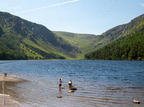 Two children play near the edge of a serene lake with a mountainous backdrop under a clear blue sky. The scene is peaceful and captures the beauty of nature.