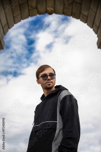 Handsome man posing for advertisement wears round glasses, looking to the camera, isolated on white blue sky  outdoor. People emotion concept