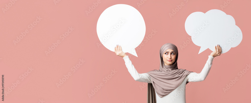 Difficult choice. Concerned arabian woman in hijab holding and choosing between two empty speech bubbles, pink studio background with free space Difficult choice. Concerned arabian woman in hijab holding and choosing between two empty speech bubbles, pink studio background with free space