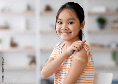 Healthy asian kid girl got vaccinated, sitting on couch at home and showing adhesive band on her shoulder, smiling at camera, panorama with copy space. Kids vaccination, immunization concept