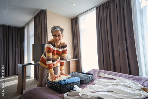 Positive senior woman packing clothes into suitcase in hotel room